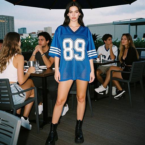 Photograph of a dark-haired woman in a blue NFL jersey number 89, black boots, standing on a rooftop terrace with seated friends. Urban skyline