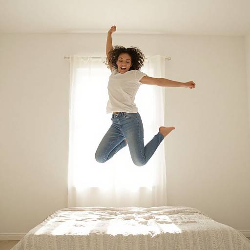 Photograph of a joyful African-American woman with curly hair, wearing a white t-shirt and blue jeans, jumping on a bed in a bright, white