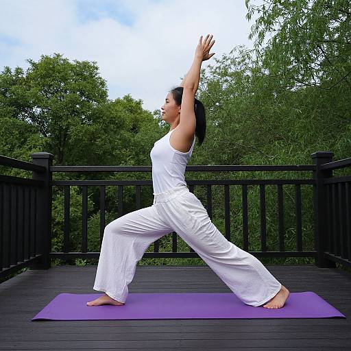 Photograph of a woman in white yoga pants and tank top, performing a tree pose on a purple mat on a black wooden deck surrounded by green trees