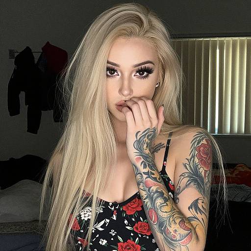 Photograph of a blonde woman with long hair, heavy makeup, floral dress, and colorful sleeve tattoos, hand near mouth, indoors.