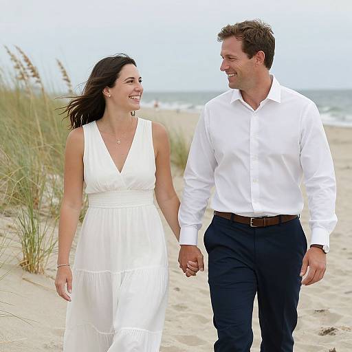 Photograph of a smiling couple holding hands on a sandy beach; woman in white sleeveless dress, man in white shirt and black pants.