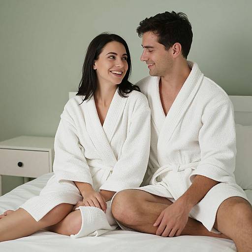Photograph of a smiling couple in white bathrobes sitting on a bed, both looking at each other, with a white nightstand in the background