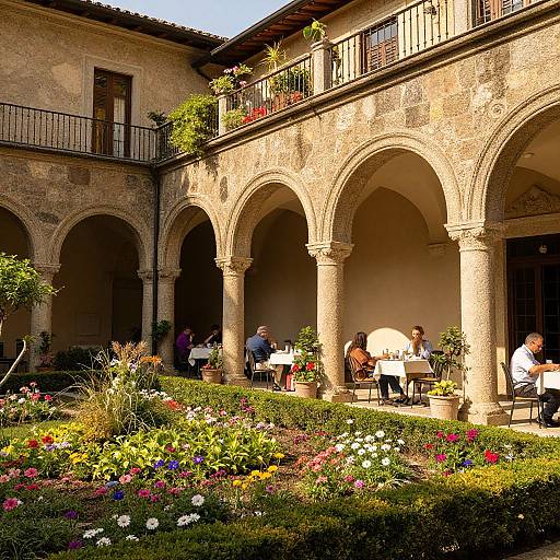 Photograph of a sunlit, rustic courtyard with arched columns, vibrant flower garden, and people dining on the shaded patio.