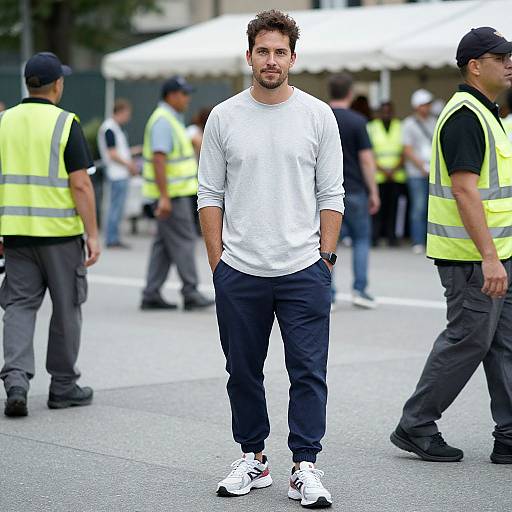 Photograph of a bearded man in a white long-sleeve shirt and navy pants, standing confidently among security personnel in neon vests. Urban street
