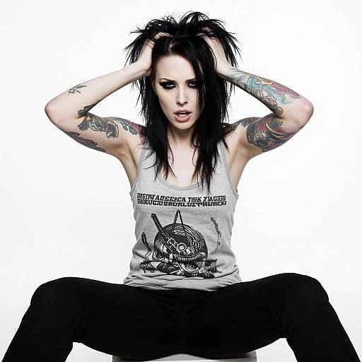 Photograph of a tattooed, pale-skinned woman with black hair, wearing a gray Metallica tank top and black pants, sitting with legs spread