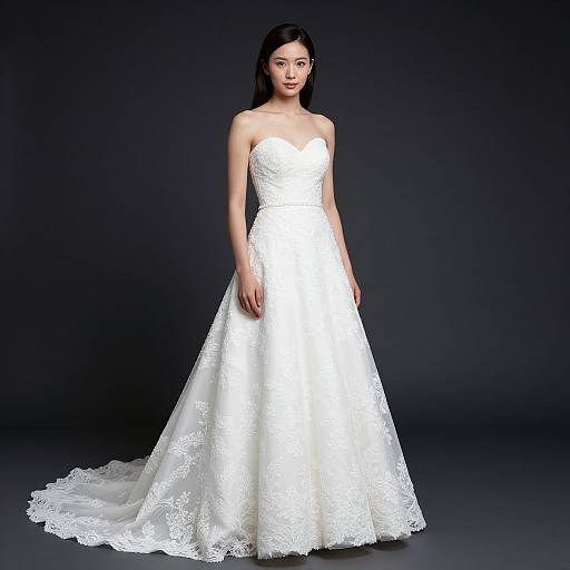 Photograph of a young Asian woman with long black hair, wearing a strapless, white lace wedding gown with a long train, standing against a dark