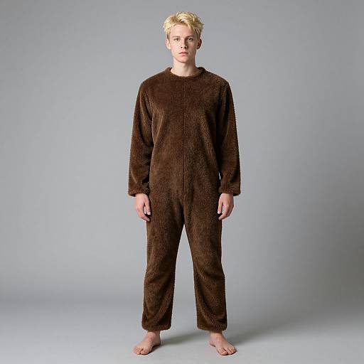 Photograph of a blonde, fair-skinned person standing barefoot against a plain gray background, wearing a loose, dark brown, fuzzy onesie.