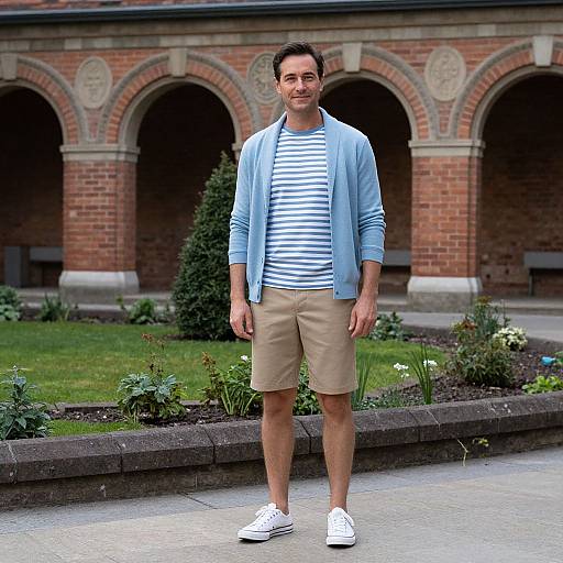 Photograph of a smiling man with short dark hair, wearing a blue cardigan, striped shirt, beige shorts, and white sneakers, standing in front