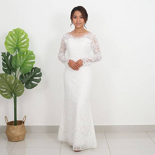 Photograph of a smiling South Asian bride in a white lace long-sleeve wedding dress, standing beside a large green monstera plant in a