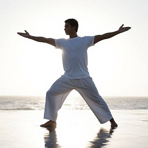 Silhouetted man in white martial arts outfit, legs wide apart, arms outstretched, standing barefoot on reflective wet sand at sunset.