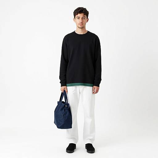 Photograph of a young man with short dark hair, wearing a black long-sleeve shirt, white pants, and black shoes, holding a navy