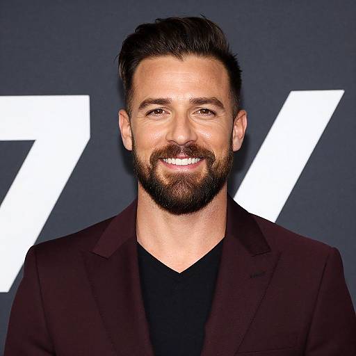 Photograph of a smiling man with dark brown hair, full beard, wearing a black shirt and dark brown blazer, against a dark blue background with