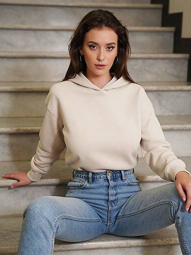 Photograph of a young woman with fair skin and brown hair, wearing a white hoodie and high-waisted blue jeans, sitting on gray stone steps