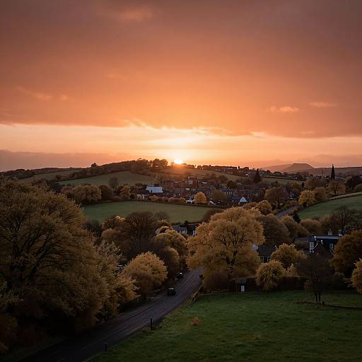 Photograph of a rural sunset with an orange-pink sky, golden autumn trees, and a small village nestled in rolling hills.