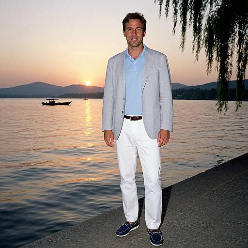 Photograph of a smiling man in a light blue blazer, white pants, and navy shoes, standing by a sunset-lit lake with a boat