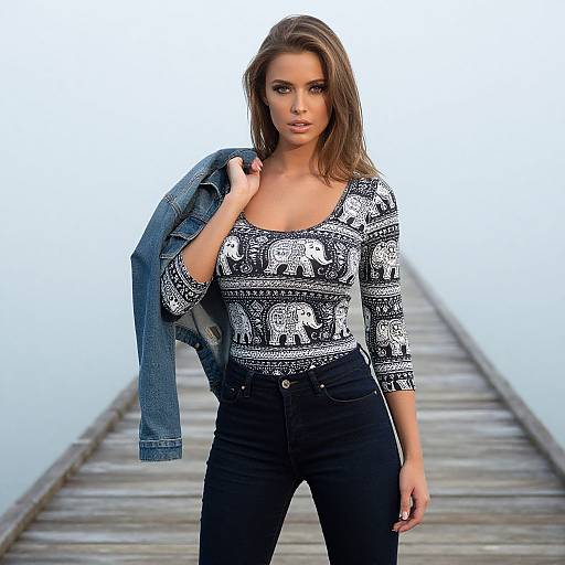 Photograph of a brunette woman with straight hair, wearing a black and white patterned blouse, black pants, and a denim jacket, standing on a