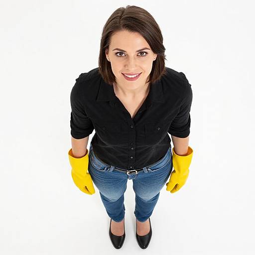 Top-down photograph of a smiling woman with medium-length dark brown hair, wearing a black button-up shirt, blue jeans, yellow gloves, and black shoes