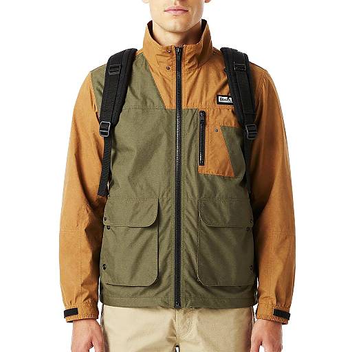 Photograph of a man wearing a green and orange outdoor jacket with black backpack, beige pants, white background. Face partially visible.