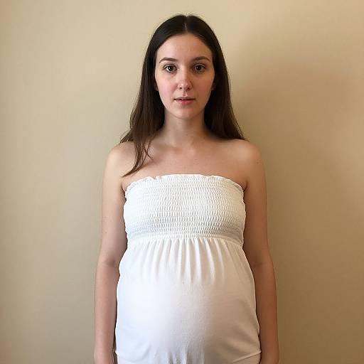 Photograph of a pregnant woman with long dark hair, wearing a strapless white dress, standing against a plain beige wall.