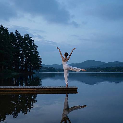 Photograph of a woman in white ballet attire, balancing on one leg with arms raised, standing on a wooden dock by a serene lake at dusk,