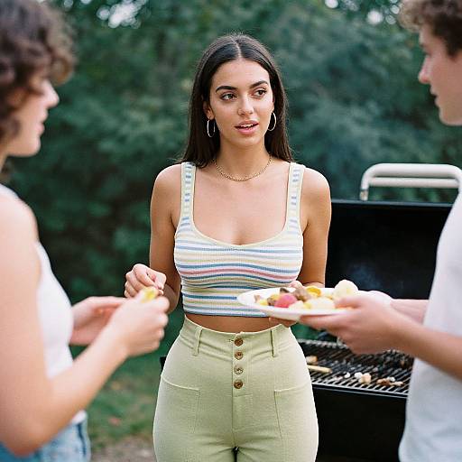 Photograph of a young woman with long black hair, wearing a striped tank top and high-waisted yellow pants, standing outdoors by a grill,