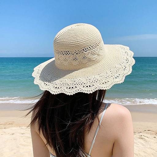 Photograph of a woman with long dark hair, wearing a white crocheted sunhat, facing a blue ocean and sandy beach.