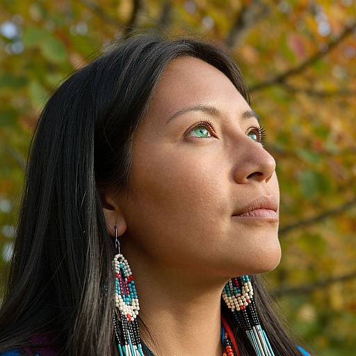 Photograph of a woman with long black hair, light brown skin, and green eyes, wearing colorful beaded earrings, gazing upward into autumn foliage