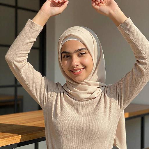 Photograph of a smiling young woman with light brown skin, wearing a white hijab and beige long-sleeve top, raising her arms joyfully