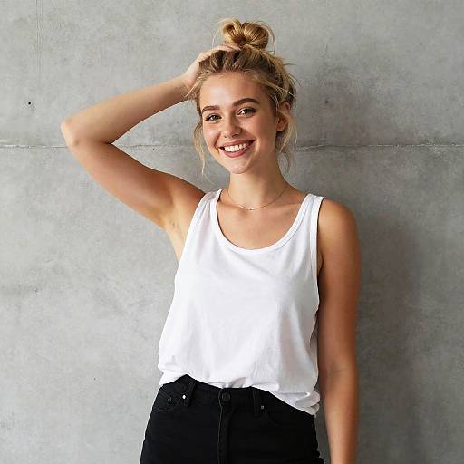 Photograph of a smiling blonde woman with a bun, wearing a white tank top and black pants, standing against a gray concrete wall.