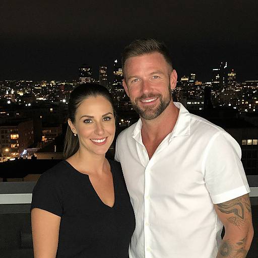 Photograph of a smiling couple on a rooftop at night, with city lights in the background. She wears a black top, he in a white shirt