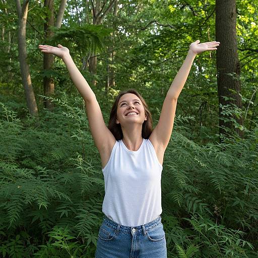 Photograph of a smiling woman with brown hair, wearing a white sleeveless top and blue jeans, raising both arms joyfully in a lush, green
