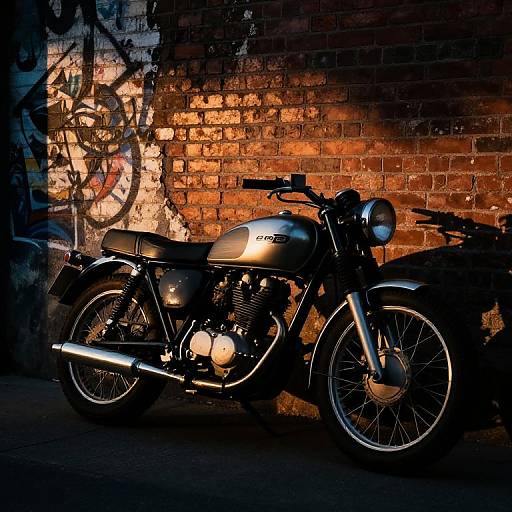 Photograph of a vintage black Honda motorcycle with glowing headlights, parked against a dimly lit, graffiti-covered, brick wall. Warm orange light highlights the
