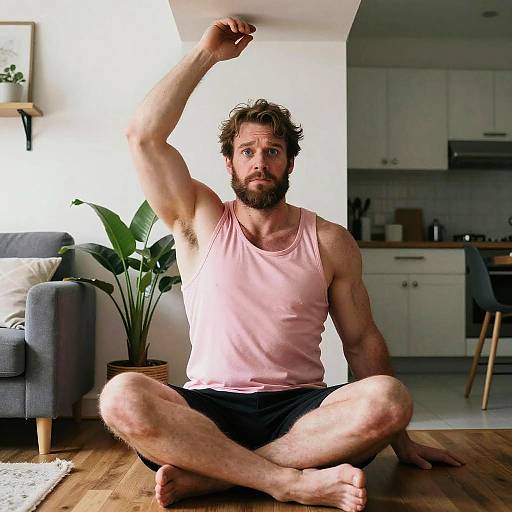 Muscular bearded man in pink tank top and black shorts, sitting cross-legged on wooden floor, stretching one arm, in modern living room.