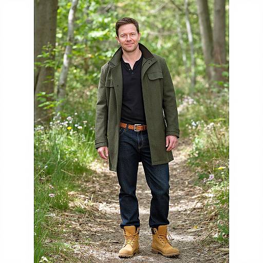 Photograph of a smiling, bearded man in a green coat, black shirt, blue jeans, and tan boots standing on a forest path.
