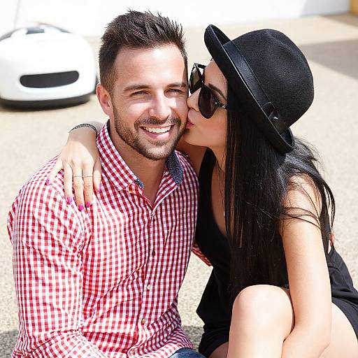 Photograph of a smiling, bearded man in a red checkered shirt, kissed on the cheek by a woman in a black hat and sunglasses.