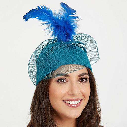 Photograph of a smiling woman with dark brown hair wearing a blue feathered hat with mesh, white background. Bright blue feathers on top.