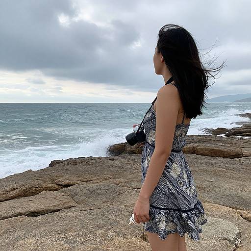 Photograph of a woman with long black hair in a patterned blue and white dress, standing on rocky coastal terrain, facing the ocean under a cloudy