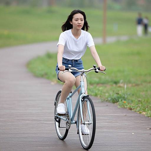 Asian woman with black hair, wearing white shirt, denim shorts, and white sneakers, riding a light blue bicycle on a park path.