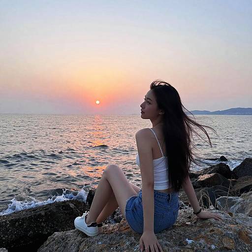 Young woman with long dark hair, white tank top, denim shorts, and white sneakers sits on rocky shore, gazing at a colorful sunset over the