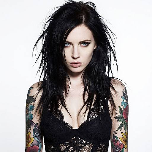 Photograph of a pale-skinned, blue-eyed woman with black, messy hair, wearing a black lace top, showing colorful tattoo sleeves, against a