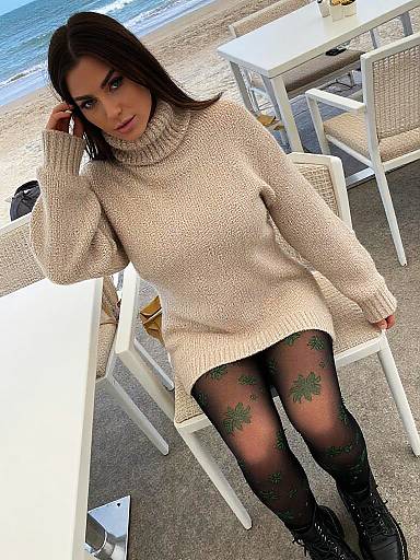 Photograph of a young woman with long brown hair, wearing a beige knit sweater, black tights with green floral patterns, seated at a beachside