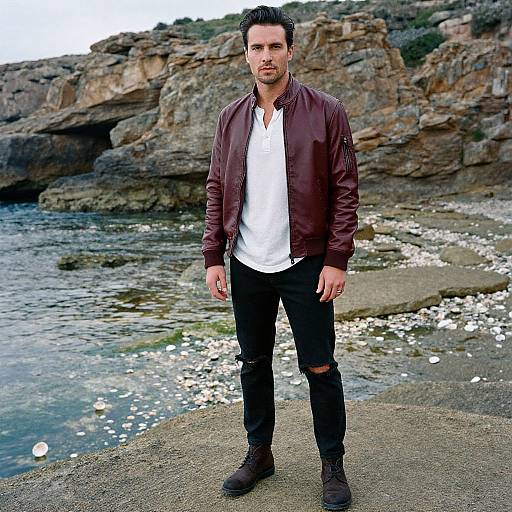 Photograph of a handsome man with short dark hair, wearing a brown leather jacket, white shirt, black ripped jeans, and boots, standing on a
