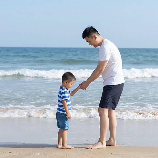 Photograph of an Asian man in a white t-shirt and black shorts, holding hands with a young boy in blue stripes, on a sunny beach with