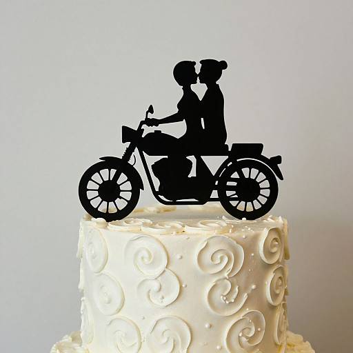 Photograph of a white wedding cake with intricate swirls, topped with a black silhouette of a couple riding a motorcycle.