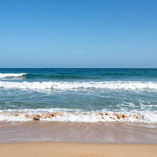 Photograph of a serene beach with clear blue sky, deep blue ocean, white foamy waves, and golden sandy shore.