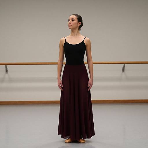 Photograph of a slender woman with dark hair in a bun, wearing a black tank top and long brown skirt, standing in a minimalist dance studio with