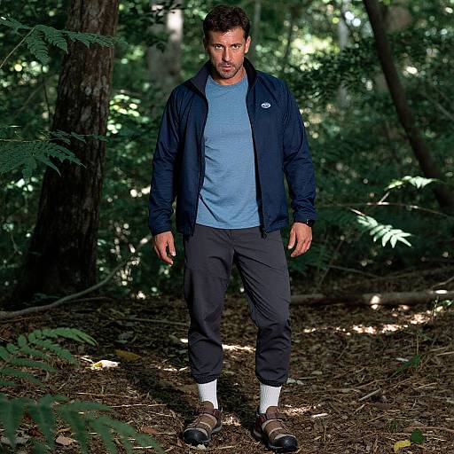 Photograph of a bearded man with short dark hair, wearing a navy jacket, light blue shirt, black pants, white socks, and brown boots