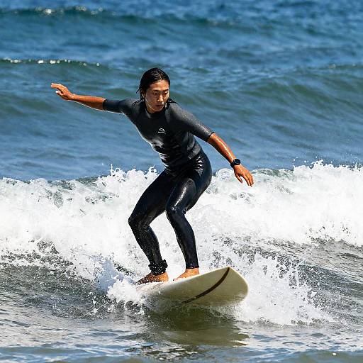 Photograph of a young man with long black hair, wearing a black wetsuit, surfing on a white surfboard in the blue ocean, with