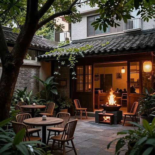 Photograph of a cozy outdoor patio with a lit fire pit, wooden chairs and tables, lush greenery, and warm indoor lights.
