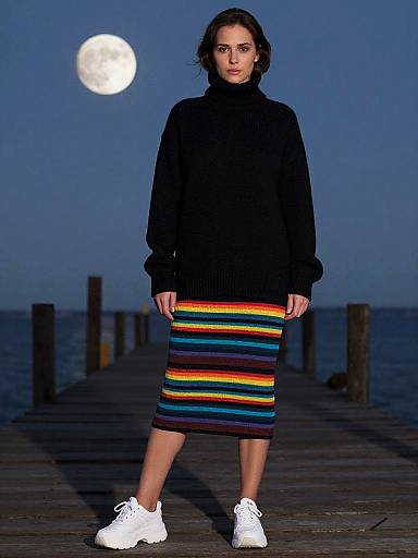 Photograph of a woman with dark hair, wearing a black turtleneck and rainbow-striped skirt, white sneakers, standing on a wooden pier at night
